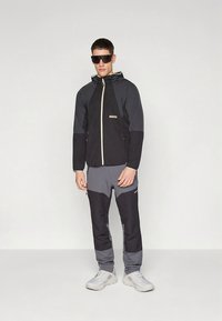 Μαύρο και γκρι μπουφάν με κουκούλα, φερμουάρ μπροστά και λογότυπο. Συνδυάζεται με γκρι παντελόνι σετ με αντίθετα πάνελ και εφαρμοστή σχεδίαση.