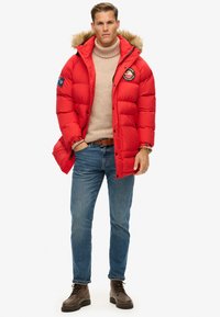 Rød puffjakke med struktureret overflade og pelsforet hætte. Har et patch på brystet og knapper langs lynlåsen. Bæres over en beige sweater.