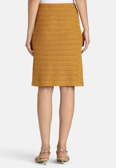 Vrouw draagt een knie-lange mosterdgele gehaakte rok en gouden sandalen met lage hak, staand met haar rug naar de camera.