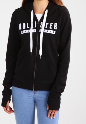 Sort hættetrøje med lynlås med hvide snore og teksten "Hollister California" båret med lyseblå jeans af en person.