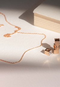 Ensemble de bijoux en or rose comprenant un collier à chaîne délicat et trois charms en forme de cubes avec des lettres gravées et un détail en pierre précieuse transparente.