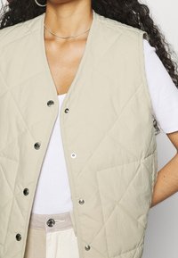 Beige gesteppte ärmellose Weste mit Druckknöpfen und Rautenmuster, getragen über einem weißen T-Shirt, kombiniert mit einem hellen Taillenbund.