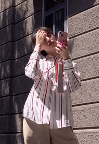 Camisa blanca con rayas verticales rojas, pantalones beige, sosteniendo un teléfono rosa con una borla. La luz del sol proyecta sombras sobre una pared texturizada.