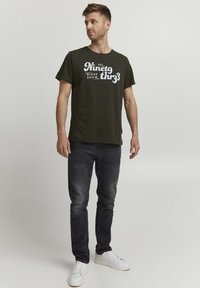 Olivengrønn T-skjorte med hvit trykt tekst "Etablert 1993 Blend Denim." Kombinert med mørke jeans og hvite joggesko. Standard passform, korte ermer.