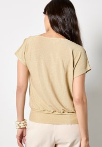 Złota bluzka z krótkim rękawem o luźnym kroju i elastycznym hemie. Błyszczący, teksturowany materiał z subtelnym, błyszczącym wykończeniem. Gładki dekolt.