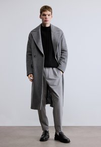 Hosszú szürke gyapjú kabát széles hajtókával, dupla soros kialakítással. Fekete bordázott pulóverrel és szürke nadrággal párosítva, fekete cipővel kiegészítve.