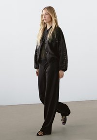 Černá oversize bunda s uzávěrem na knoflík, černý top a široké kalhoty. Kombinováno s sandály na řemínky a vlajícími vlasy. Hladké textury po celém outfitu.