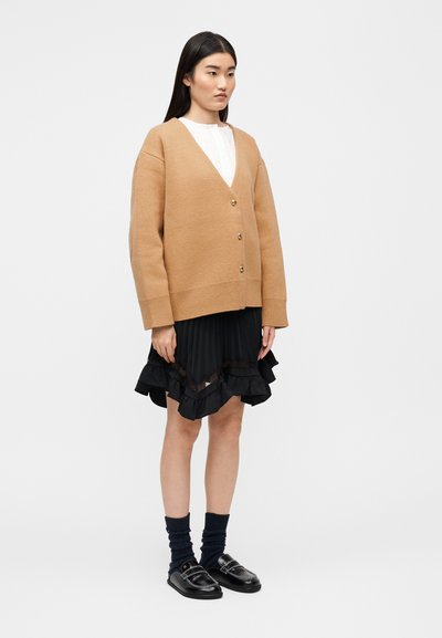 Ζακέτα από πλεκτό ύφασμα σε καμηλό χρώμα με V-λαιμόκοψη, κουμπιά μπροστά και ανοιχτή εφαρμογή, συνδυασμένη με μαύρη πλισέ φούστα με βολάν και μαύρα παπούτσια με ασημένιες λεπτομέρειες.