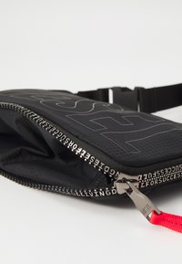Black zippered pouch made of textured fabric with a prominent logo, featuring silver hardware and a red strap accent.