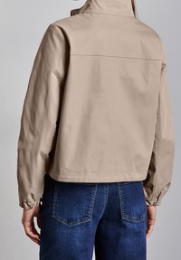 Veste beige courte avec col montant, poignets élastiques et texture lisse. Associée à un jean en denim bleu foncé.