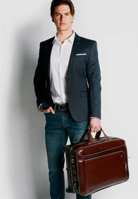 Braune Lederaktentasche mit doppelten Griffen, rechteckiger Form, Reißverschluss und vordere Tasche. Getragen von einer Person mit einem Anzug und Jeans.