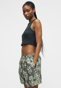 Γυναίκα με πλεξούδες, που φοράει μαύρο αμάνικο crop top και γκρι καρό σορτς με λουλούδια, με τα χέρια στις τσέπες, στραμμένη πλάγια μπροστά σε ένα απλό φόντο.