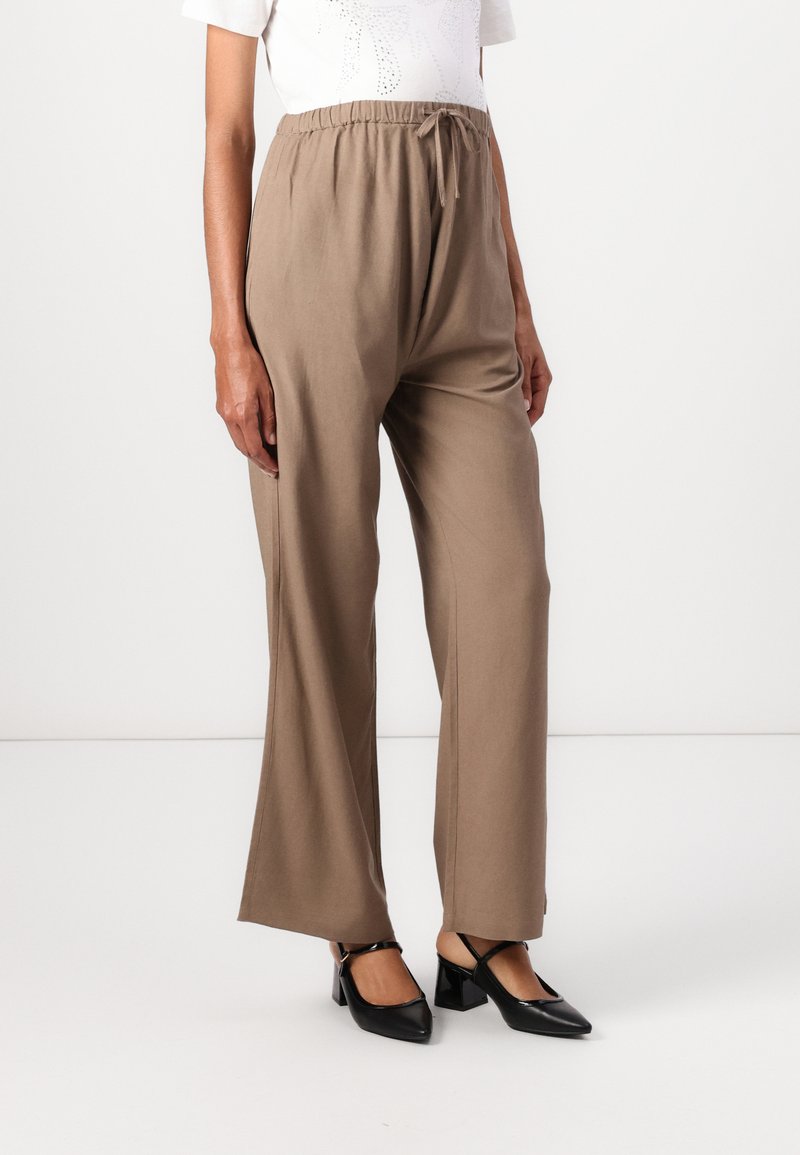 Pantalons marron à jambes larges avec taille élastique et cordon de serrage. Associés à des chaussures à talons noires, le tout sur un fond neutre.