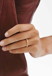 Anillo texturizado en oro que se lleva en una mano, sosteniendo un objeto de cuero burdeos. El anillo presenta una superficie lisa y pulida, y un diseño delgado.