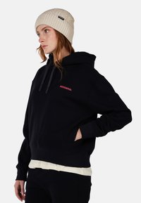 Felpa nera con cappuccio e tasca frontale, caratterizzata da un logo rosso "ROSSIGNOL". Indossata con un berretto lavorato a maglia color crema. Tessuto liscio, vestibilità comoda.