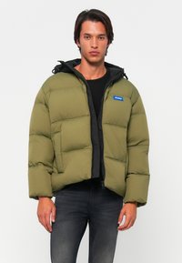 Olive green puffer jacket with a black hood, featuring a zip front and a blue logo patch, designed with a boxy shape and quilted texture.