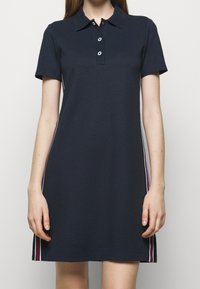 Mörkblå polo-klänning med en texturerad tyg, korta ärmar och spetsad krage; har vita och röda sidostripes och treknapplåst.