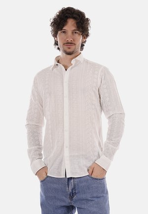 Hombre joven con cabello rizado y oscuro, vestido con una camisa blanca de botones con estampado y jeans azules, de pie con las manos en los bolsillos sobre un fondo blanco.