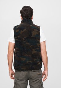 Fleece kamufliažo liemenė su aukštu apykakle, turinti tamsiai žalios ir rudos spalvos raštų mišinį, vilkima ant baltos marškinėlių.