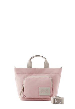 Rosa Tragetasche mit einer vorderen Reißverschlusstasche mit der Aufschrift "Studio Berlin", beigen Stoffgriffen und abnehmbarem beigen Schultergurt daneben gelegt.