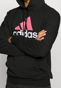 Felpa nera con cappuccio caratterizzata da un logo Adidas rosa e bianco. Realizzata in tessuto morbido, con tasca a marsupio e cappuccio regolabile con cordini.
