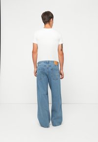 Mann som står med ryggen mot, iført en hvit t-skjorte og løstsittende blå jeans med baklommer og en tatovering på høyre arm.