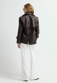Giacca in finta pelle marrone con schiena drappeggiata e polsini con cintura, abbinata a pantaloni bianchi a gamba larga. Tessuto liscio e vestibilità sartoriale.