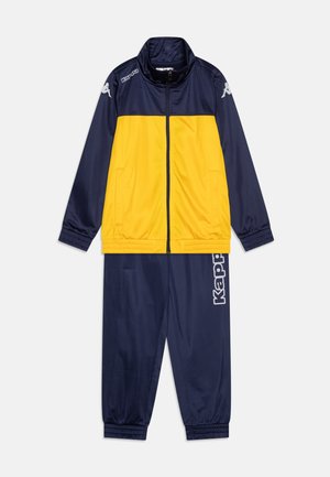Survêtement bleu marine et jaune avec une veste zippée, comportant des poches latérales et des poignets élastiques, ainsi que des pantalons assortis avec un logo imprimé sur le côté.