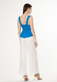 Blauwe peplumtop met brede bandjes en gerimpelde achterkant, gecombineerd met witte wijde broek en goudkleurige hakken.