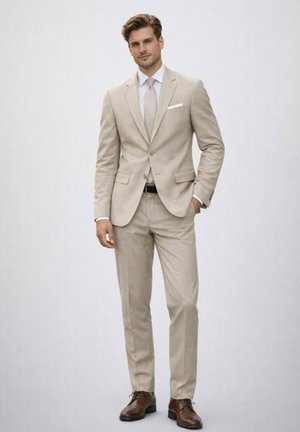 Homme debout portant un costume beige avec une cravate assortie, une chemise blanche, des chaussures de ville marron et une main dans la poche, devant un fond uni.
