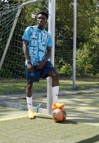 Tricou sportiv albastru cu modele geometrice, asortat cu pantaloni scurți bleumarin. Minge de fotbal portocalie la picioare, ghete portocalii și șosete albe. Fundal cu un teren verde.