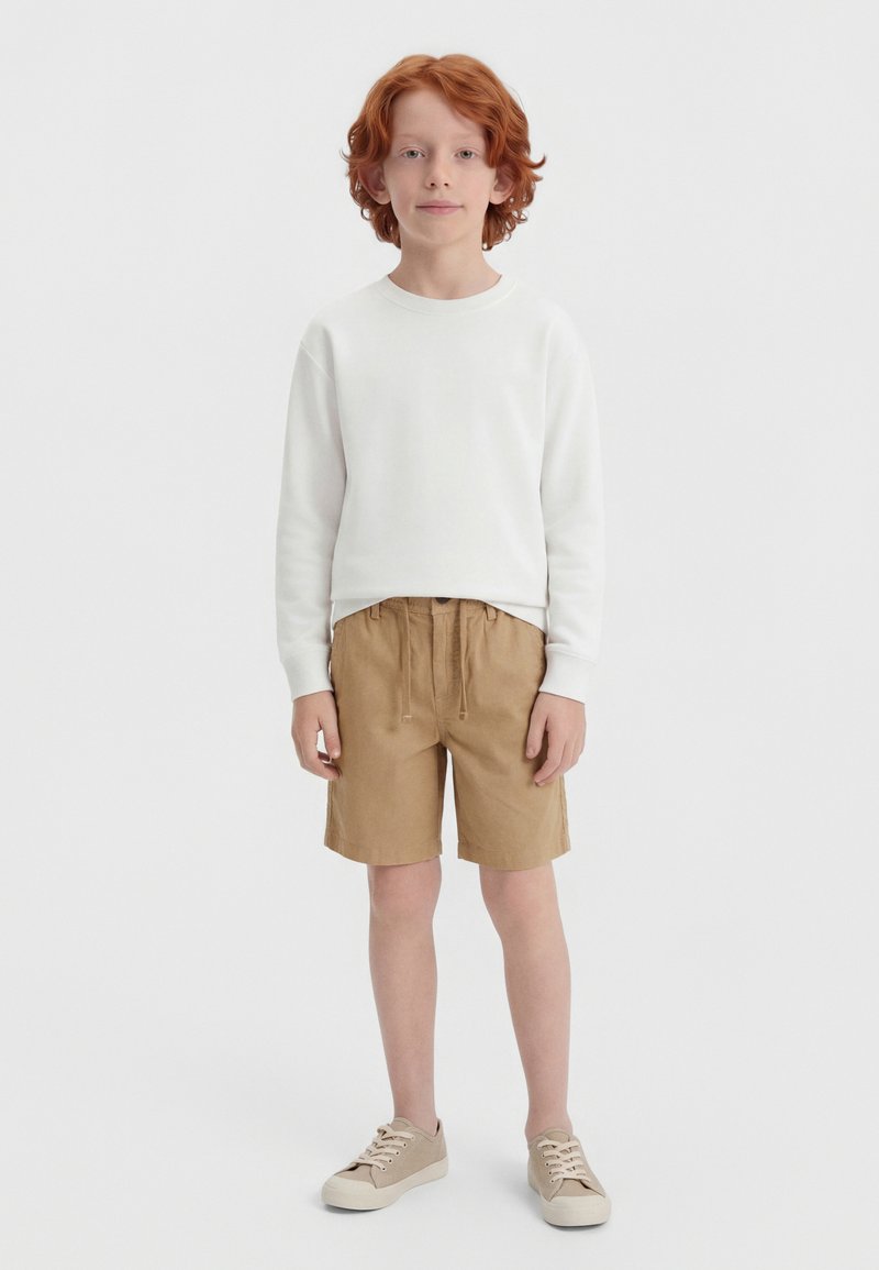 Παιδί με κόκκινα μαλλιά στέκεται φορώντας λευκό μακρυμάνικο πουκάμισο, μπεζ σορτς και μπεζ αθλητικά παπούτσια μπροστά σε απλό φωτεινό φόντο.