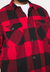 Camicia di flanella a quadri rossi e neri con due tasche sul petto dotate di bottoni neri e colletto classico. Il tessuto ha un aspetto texture.