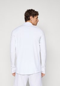 Mann med krøllete hår iført langermet hvit skjorte og hvite shorts, stående med ryggen til mot en enkel lys bakgrunn.