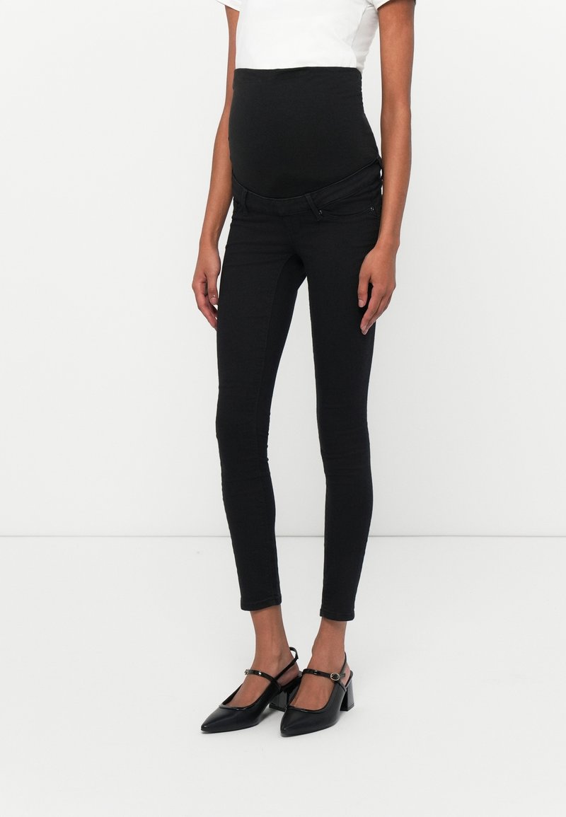 Vaqueros skinny de maternidad negros con una textura suave, cintura alta y bolsillos frontales. Combinados con zapatos de tacón negros.