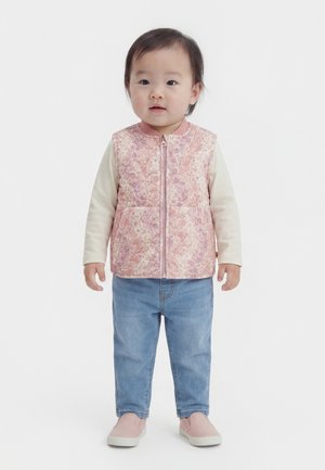 Kisgyerek áll rózsaszín virágos steppelt mellényben, krémszínű hosszú ujjú ingben, kék farmerben és rózsaszín bebújós cipőben, fehér háttér előtt.