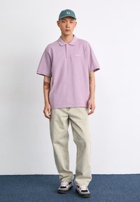 Chemise polo lavande avec un col à trois boutons, associée à un pantalon large beige clair et des baskets noires et roses, sur fond uni.