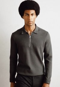 Pulover tricotat de culoare gri închis, cu guler polo cu fermoar, mâneci lungi și o textură netedă, asortat cu pantaloni negri. Design minimalist.