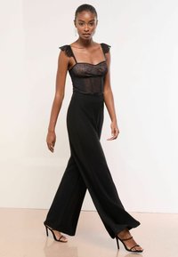 Pantalones anchos negros combinados con un body negro ajustado que presenta un intrincado detalle de encaje y tirantes con volantes; unos tacones de aguja completan el look.