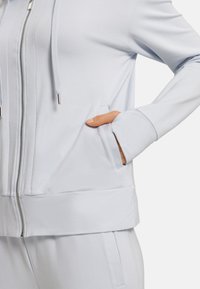 Jasnoniebieska bluza zapinana na zamek, wykonana z miękkiego materiału, z kapturem, kieszeniami i lekko dopasowanym pasem, ozdobiona przeszyciami.