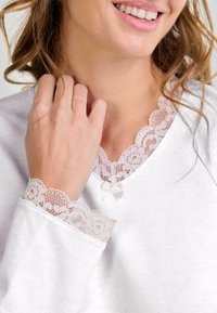 Mujer sonriendo suavemente, vestida con una blusa blanca con encaje en el cuello y el puño, sosteniendo un mechón de su pelo rizado cerca de su cara.