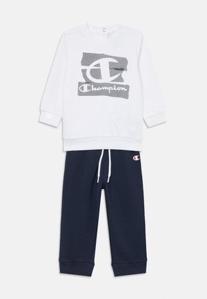 Balta džemperis ar pelēku Champion logotipu, garām piedurknēm un ribotu aproci; saskaņots ar tumši zilo sporta biksēm, kurām ir logotipa ielāps un aukla.