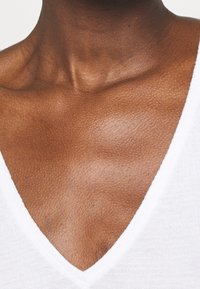 Top blanco de tela con escote en V que muestra una piel suave y texturizada y clavículas pronunciadas, con sutiles brillos en la superficie de la piel.
