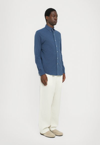 Chemise bleu à boutons avec un tissu texturé, associée à un pantalon ample de couleur crème et des chaussures beiges, se tenant devant un fond blanc.