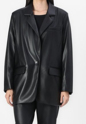 Osoba ubrana w czarną marynarkę z imitacji skóry z jednym guzikiem, kołnierzem z wycięciem, klapami kieszeni oraz dopasowanymi spodniami na jednolitym tle.