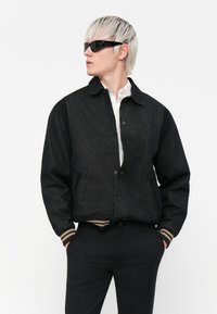 Svart denim bomberjakke med kontrasterende svarte ermer, knappelukking og ribbestrikket striped hem. Båret over en hvit skjorte, sammen med mørke bukser.