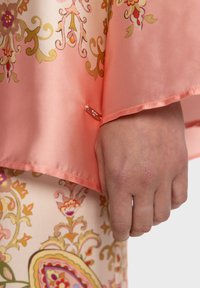 Blouse en soie de couleur corail, ornée de motifs floraux et cachemire en or et rose, détaillée avec un accent en métal doré sur le poignet.