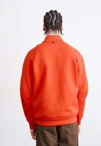 Sweatshirt laranja com gola alta e punhos canelados, apresentando um pequeno detalhe circular na parte de trás e um design de corte descontraído.