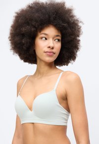 Γυναίκα με πλούσια σγουρά μαλλιά που φοράει ανοιχτόχρωμο, ριμπ, σουτιέν με V-λαιμόκοψη, κοιτάζοντας προς τα αριστερά της, σε απλό λευκό φόντο.