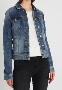 Denim jacket in medium blue with a slightly distressed finish, featuring copper buttons, two chest pockets, and buttoned cuffs.
